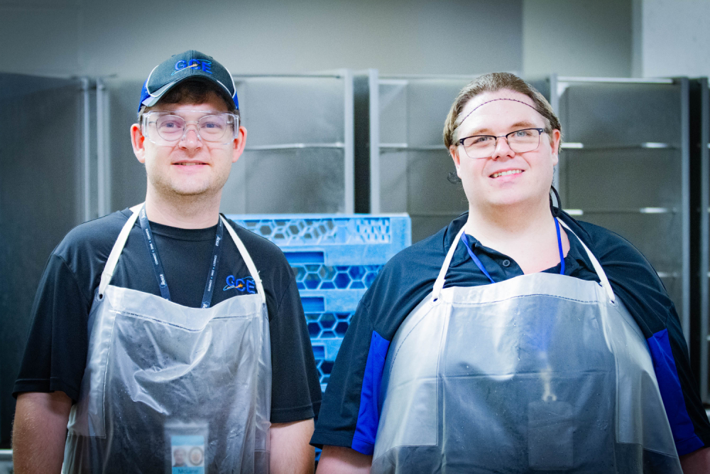 Two male coworkers standing side-by-side wearing aprons and smiling