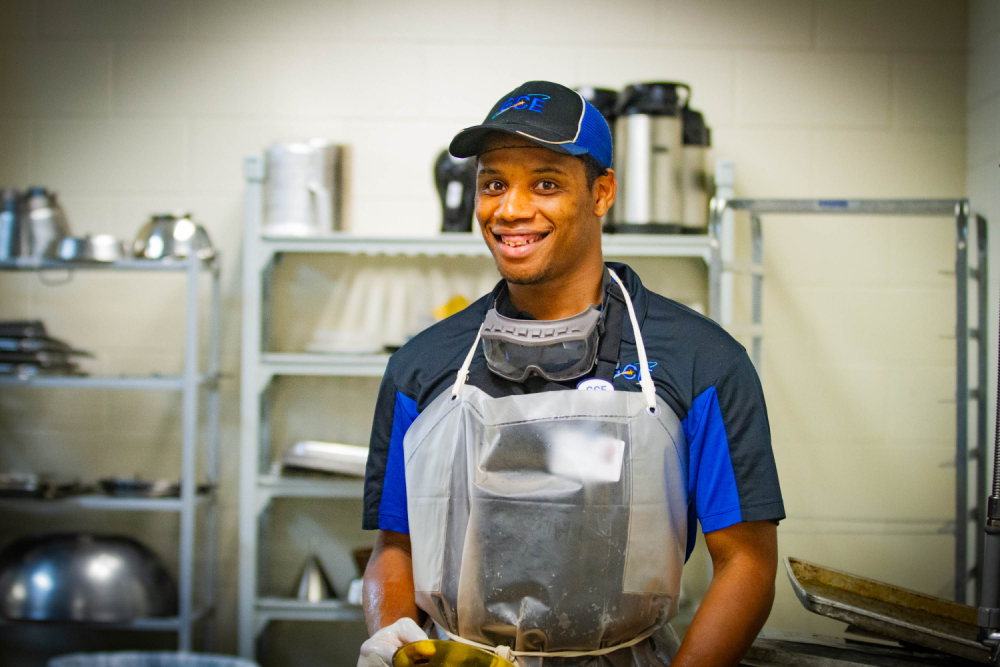 Man in kitchen wearing apron and GCE cap smiling